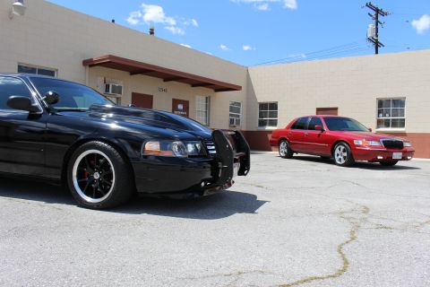 2003 Ford Crown Victoria - black Ford Crown Victoria with a push bar sits in the foreground while a red Mercury Grand Marquis is parked across the industrial lot unde…