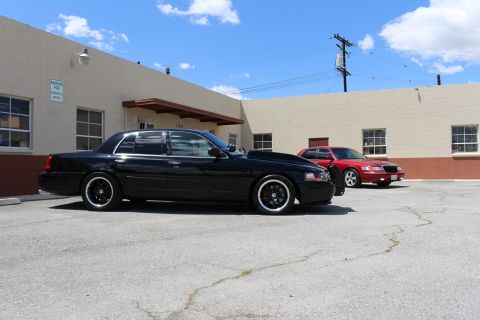 2003 Ford Crown Victoria - Side profile of a lowered black Ford Crown Victoria on black wheels parked beside an industrial building, with a red Mercury sedan in the b…