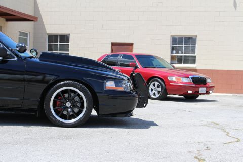 2003 Ford Crown Victoria - Low front-fender view of a black Ford Crown Victoria on black wheels with a red Mercury Grand Marquis parked beside it at an industrial bui…