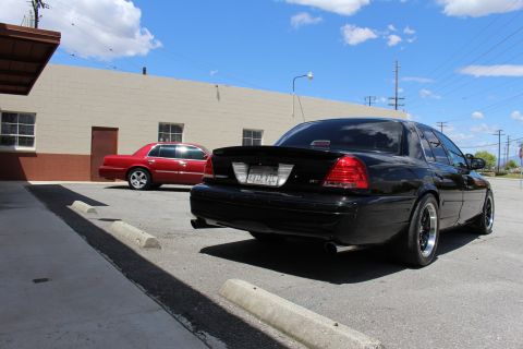 2003 Ford Crown Victoria - Rear three-quarter view of a lowered black Ford Crown Victoria with a trunk spoiler and dual-tip exhaust, a red sedan parked in the backgro…