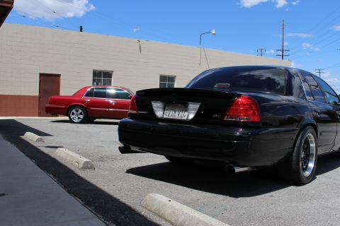2003 Ford Crown Victoria - Rear three-quarter view of a black Ford Crown Victoria with a spoiler and 'EVIL VIC' plate, with a red Mercury sedan parked behind in the l…