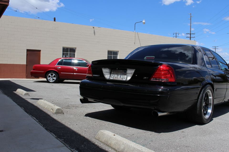 Rear three-quarter of a black sedan with spoiler and EVIL VIC plate, red car behind