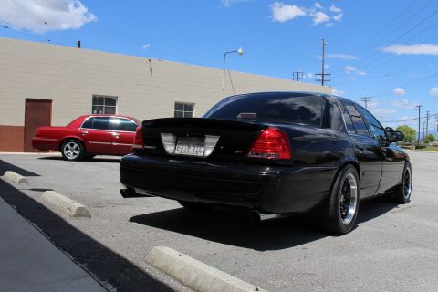 2003 Ford Crown Victoria - Rear three-quarter view of a black Ford Crown Victoria with a trunk spoiler and dual exhaust, a red Mercury sedan parked across the industr…