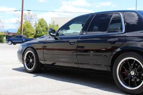 2003 Ford Crown Victoria - Side profile of a lowered black Ford Crown Victoria on polished-lip black wheels in a paved lot, with a dark pickup truck parked in the bac…