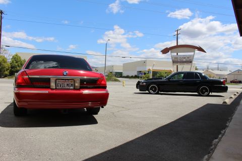 2003 Ford Crown Victoria - Red Mercury sedan rear with chrome trim and EVIL MGM plate, parked near a lowered black sedan beneath a curved retro sign.