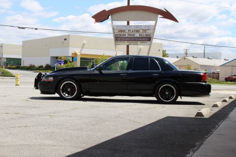 2003 Ford Crown Victoria - lowered black full-size sedan shown in side profile with dark wheels, parked beneath a curved retro sign at an industrial lot.