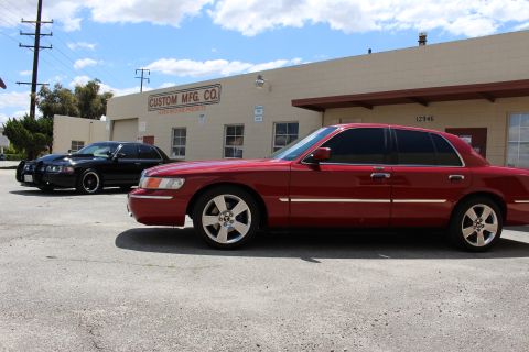 2003 Ford Crown Victoria - Side profile of a red Mercury sedan with chrome trim and a black sedan parked behind it outside the Custom Mfg Co building.
