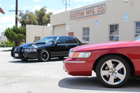 2003 Ford Crown Victoria - Close view of a red sedan's headlight and machined wheel in the foreground, with a black sedan parked by the Custom Mfg Co building.