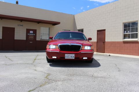 2003 Ford Crown Victoria - Front view of a red Mercury sedan with its headlights illuminated and a partial EVIL MGM plate, parked between two industrial buildings.