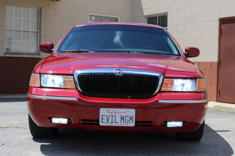 2003 Ford Crown Victoria - Close front shot of a red Mercury sedan showing the Mercury emblem, chrome grille, lit headlights, and California EVIL MGM plate.