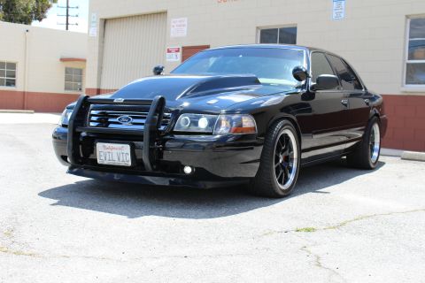 2003 Ford Crown Victoria - Black Ford Crown Victoria sedan with a brush guard and California EVIL VIC plate, parked beside the Custom Mfg building.