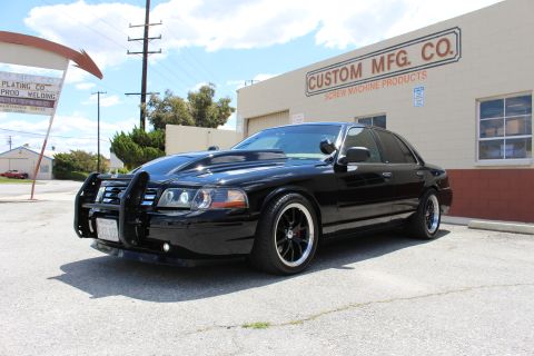 2003 Ford Crown Victoria - Black Ford Crown Victoria with a chrome grille guard and dark wheels, parked in front of the Custom Mfg Co building under blue sky.