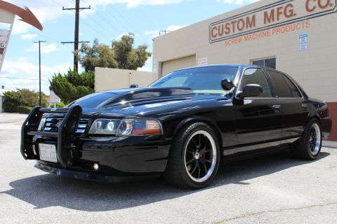 2003 Ford Crown Victoria - black full-size Ford sedan fitted with a front push bar and dark aftermarket wheels sits in a parking lot outside an industrial building.