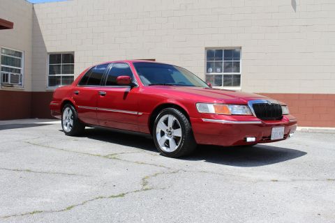 2003 Ford Crown Victoria - red Mercury full-size sedan with chrome trim and alloy wheels is parked on cracked pavement beside a tan building with windows.