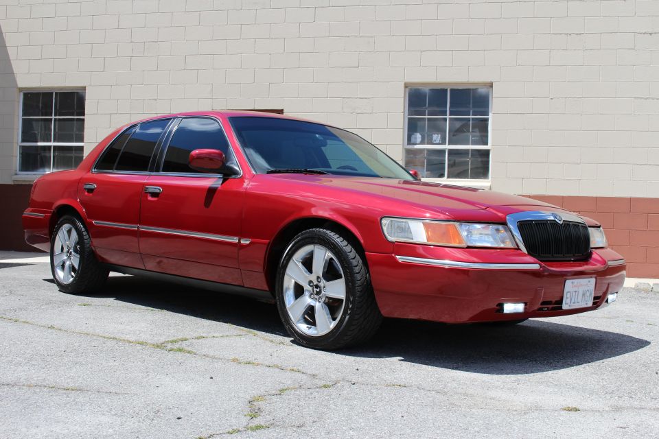 Red Mercury Grand Marquis sedan, front three-quarter view, by tan building