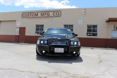2003 Ford Crown Victoria - Head-on view of a black Ford sedan with a chrome grille guard and a license plate reading EVIL VIC, parked outside an industrial building.