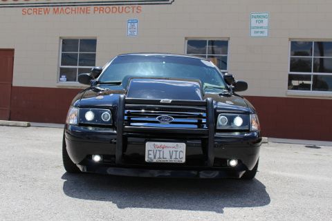 2003 Ford Crown Victoria - black Ford Crown Victoria sedan shown head-on with a Ford grille guard and a California EVIL VIC plate, parked by a tan building.