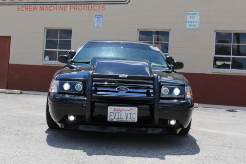 2003 Ford Crown Victoria - Head-on shot of a black Ford Crown Victoria with a chrome grille guard and California EVIL VIC plate, on pavement by a tan wall.