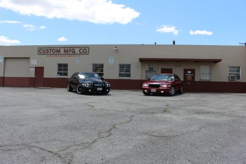 2003 Ford Crown Victoria - black sedan and a red sedan parked side by side on cracked pavement in front of a long tan industrial building under blue sky.