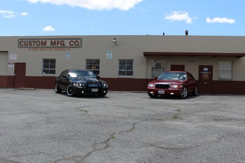 2003 Ford Crown Victoria - black Ford sedan with grille guard and a red Mercury sedan parked together in a lot outside the Custom Mfg. Co. building.