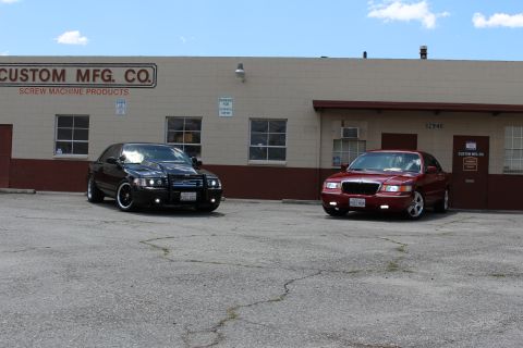2003 Ford Crown Victoria - black Ford sedan and a red Mercury sedan parked on cracked asphalt in front of a tan industrial building marked Custom Mfg. Co.