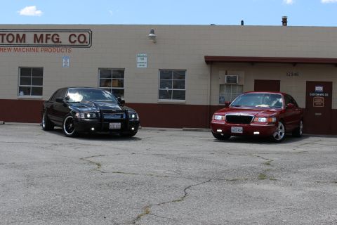 2003 Ford Crown Victoria - black Ford sedan and a red Mercury sedan parked some distance apart on cracked pavement before a tan industrial building.
