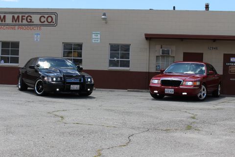2003 Ford Crown Victoria - black Ford sedan with grille guard and a red Mercury sedan parked on cracked asphalt in front of a tan industrial building.