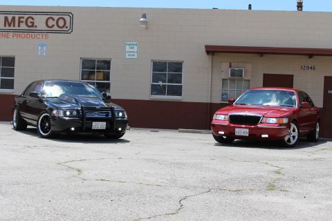 2003 Ford Crown Victoria - black Ford sedan and a red Mercury sedan parked at slight angles on cracked pavement in front of a tan industrial building.