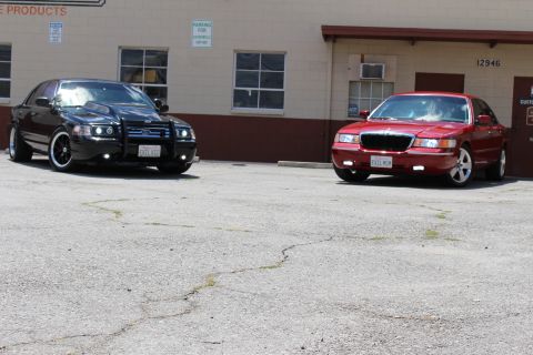 2003 Ford Crown Victoria - black Ford sedan and a red Mercury sedan, both front-facing, parked on cracked asphalt before a tan industrial building.