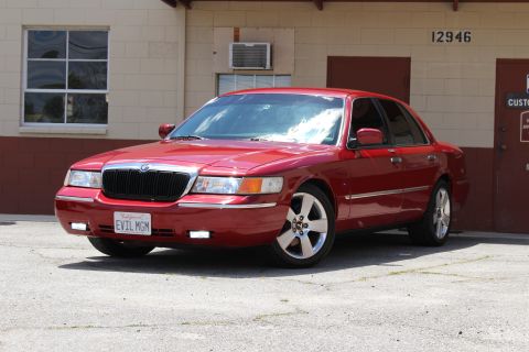 2003 Ford Crown Victoria - red Mercury full-size sedan with chrome waterfall grille and a California EVIL MGM plate, parked angled by a cinder-block building.