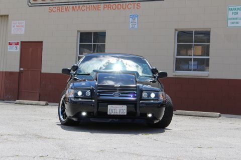 2003 Ford Crown Victoria - Head-on view of a black Ford Crown Victoria with grille guard and EVIL VIC plate, sunlight glaring off the windshield, by an industrial wal…