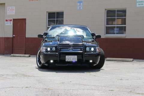 2003 Ford Crown Victoria - Head-on shot of a black Ford Crown Victoria with a Ford-badged grille guard, projector lights and EVIL VIC plate, parked by an industrial w…