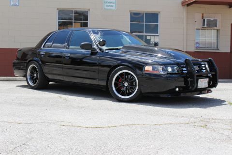 2003 Ford Crown Victoria - lowered black Ford Crown Victoria sedan with a chrome-lipped black wheels and a front grille guard parked beside a tan building.