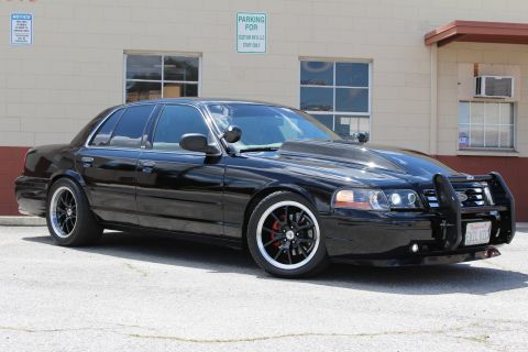 2003 Ford Crown Victoria - lowered black Ford Crown Victoria with grille guard, blacked-out trim and chrome-lip wheels parked on pavement by a tan wall.