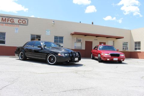 2003 Ford Crown Victoria - black Ford Crown Victoria beside a red Mercury Grand Marquis on cracked pavement outside a tan industrial building.