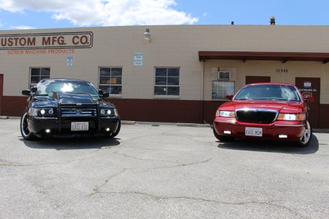 2003 Ford Crown Victoria - Front view of a black Ford Crown Victoria with halo headlights and grille guard parked next to a red Mercury sedan by a building.