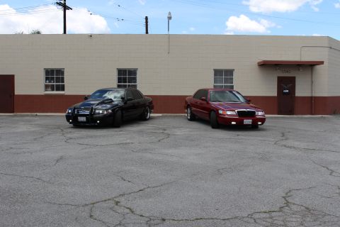 2003 Ford Crown Victoria - Wide shot of a black Ford Crown Victoria and a dark red Mercury Grand Marquis parked on cracked pavement by a tan building.