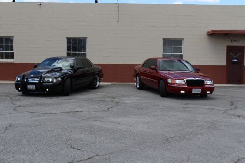 2003 Ford Crown Victoria - black Ford Crown Victoria with grille guard parked beside a dark red Mercury Grand Marquis outside an industrial building.