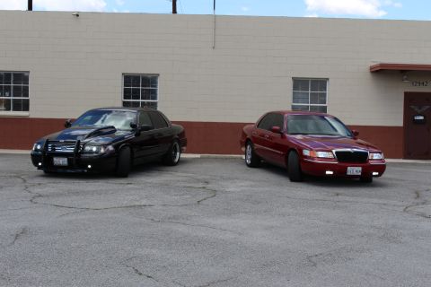 2003 Ford Crown Victoria - Front three-quarter wide view of a black Ford Crown Victoria and a red Mercury Grand Marquis parked by a tan building.