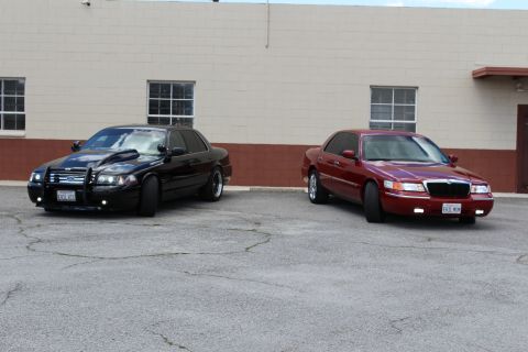 2003 Ford Crown Victoria - black Ford Crown Victoria with grille guard sits beside a dark red Mercury Grand Marquis against a tan block wall.
