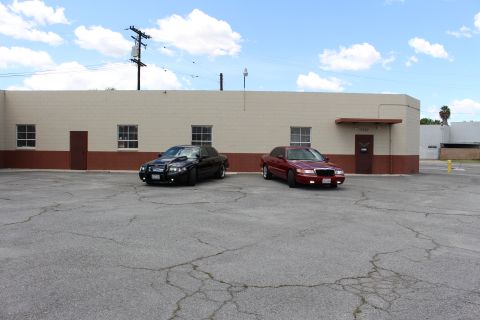2003 Ford Crown Victoria - distant wide shot of a black sedan and a dark red sedan parked on cracked pavement before a tan building under blue sky.