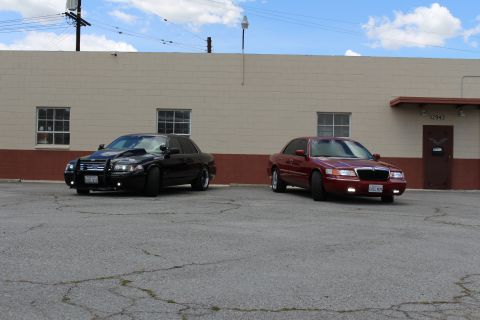2003 Ford Crown Victoria - black Ford Crown Victoria with grille guard beside a dark red Mercury Grand Marquis on pavement outside a tan building.