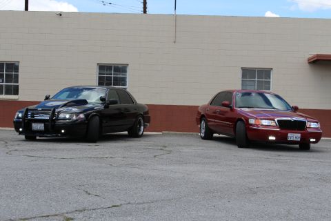 2003 Ford Crown Victoria - blacked-out sedan fitted with a police push bumper parks next to a maroon four-door sedan along a building wall.