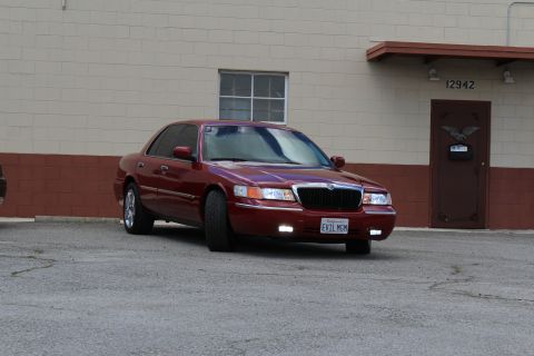 2003 Ford Crown Victoria - maroon Mercury Grand Marquis with chrome grille and California plate EVIL MOM sits angled before a tan warehouse.