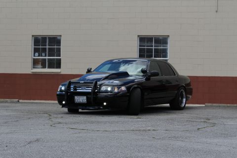 2003 Ford Crown Victoria - lowered black Ford Crown Victoria fitted with a police push bumper and aftermarket wheels sits before a tan building.
