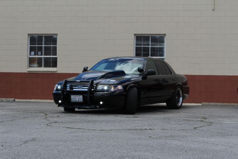 2003 Ford Crown Victoria - black Ford Crown Victoria with a police push bumper and chrome wheels poses in a lot before a paned-window wall.