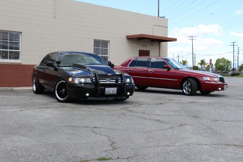 2003 Ford Crown Victoria - black Ford Crown Victoria with a push bumper parks beside a maroon Mercury sedan in a paved lot under blue sky.