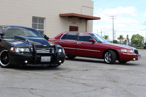 2003 Ford Crown Victoria - black Ford Crown Victoria with push bumper sits front-angled near a maroon Mercury sedan in a sunny parking lot.