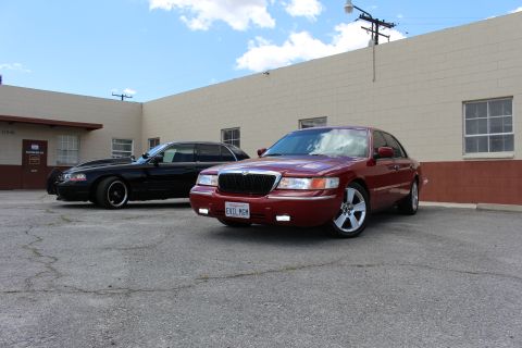 2003 Ford Crown Victoria - maroon Mercury Grand Marquis sits in the foreground with a lowered black Ford sedan behind, both in a paved lot.