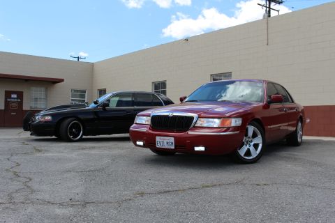 2003 Ford Crown Victoria - maroon Mercury Grand Marquis with chrome grille and plate EVIL MOM parks beside a black Ford sedan under blue sky.